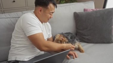 Genç bir Çinli evcil köpeğiyle evdeki iç mekanında dizüstü bilgisayarı olan rahat bir oturma odasında kanepede oynuyor..
