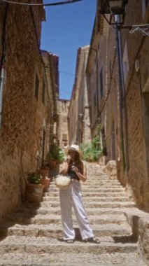 Fornalutx, Mallorca, İspanya 'da taş basamaklarda duran, şapka ve güneş gözlüğü takan, elinde telefon tutan, güneşli bir günde taş binalar ve bitkilerle çevrili bir kadın. 