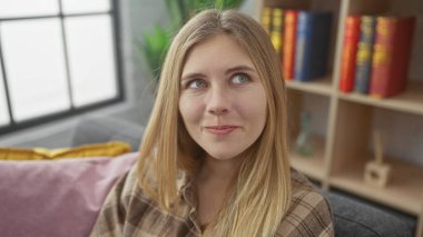 Sıradan sarışın bir kadın, modern bir oturma odasında, kitaplık arka planıyla rahatlıyor..