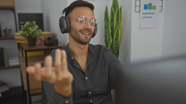 Sakallı, gözlüklü, kulaklık takan, raflarla ve bitkilerle dolu bir ofiste otururken jestler yapan genç bir İspanyol..