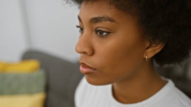 Düşünceli, kıvırcık saçlı, zarif ve sakin bir şekilde modern bir oturma odasında poz veren Afrikalı Amerikalı bir kadın..