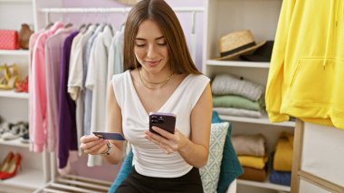 Renkli bir gardırop odasında elinde kredi kartıyla akıllı telefon kullanan bir kadın..