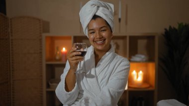 Bir spa odasında genç bir İspanyol kadın, bornoz ve havlu giyiyor, elinde bir bardak şarapla gülümsüyor, mumlarla çevrili ve huzurlu bir atmosferi var..