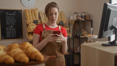Fırın tezgahında arka planda ekmek olan akıllı telefon kullanan bir kadın, iç mekandaki müşteriler için modern ve davetkar bir ortam yaratıyor.