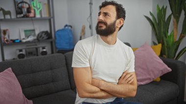 Düşünceli orta yaşlı sakallı adam modern oturma odasında oturuyor, kollarını kavuşturup düşünüyor..