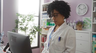 Kıvırcık saçlı Afrikalı bir Amerikalı kadın eczacı, laboratuvar önlüğü ve gözlük takan bir bilgisayar ve ürünlerle dolu bir eczanede çalışıyor..