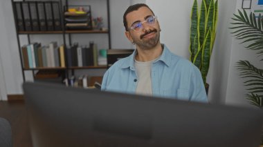 Ofis ortamında çalışan, bıyıklı yakışıklı İspanyol adam masasında otururken gülümsüyor.