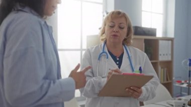 Klinikte bir hastaya danışmanlık yapan kadın bir doktor, göz teması kurarken not alır, kapalı bir klinik ortamında profesyonel bir tıbbi atmosfer yaratır..