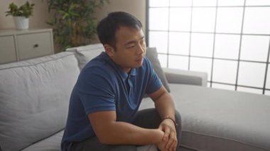 Mavi gömlekli genç, yakışıklı bir Çinli modern, bitki süslemeli bir oturma odasında gri bir kanepede ayağa kalkar..