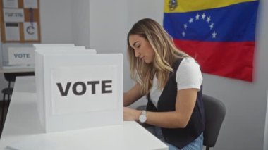 Genç, sarışın bir kadın seçim odasında oy kullanıyor. Venezuela bayrakları ve oy kullanma kabinleriyle..