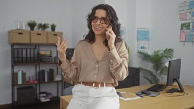 Ofis ortamında güzel bir esmer kadın, gözlük takıyor, telefonla konuşuyor arka planda kapalı bir masa başında bitkiler ve kitaplıklarla oturuyor..