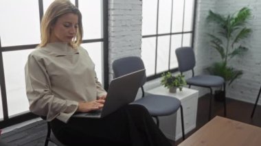 Genç ve güzel bir kadın, modern, parlak bir bekleme odasında otururken dizüstü bilgisayarı üzerinde çalışıyor. Etrafı sandalye ve bitkilerle çevrili, iyi ışıklandırılmış bir iç mekanda..