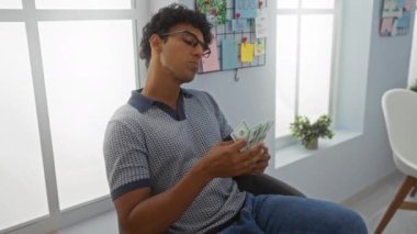 Genç adam, modern bir ofis odasında duvarında parlak dekorasyon ve yaratıcı fikir panosuyla oturmuş Amerikan dolarlarını sayıyor.