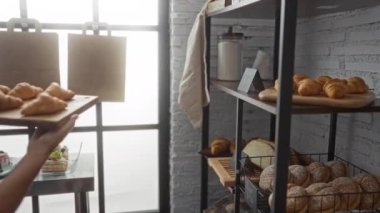 Orta yaşlı bir kadın sıcak pastanede raflara taze kruvasan yerleştiriyor. Ekmek ve pasta çeşitleri raflarda güzelce sergileniyor ve ev ortamında davetkar bir ortam yaratıyor.