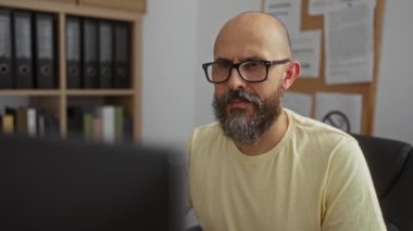 Gözlüklü ve sakallı bir adam ofis ortamında çalışıyor, arkasında belgeler ve klasörlerle bir masada oturuyor, odaklanmış ve bilgisayar ekranlarıyla meşgul görünüyor.