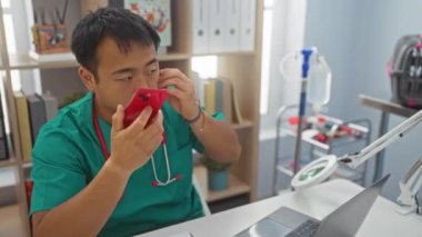 Genç Asyalı erkek veteriner, bir klinik odasında, laptopta çalışırken kırmızı akıllı telefonuna konuşuyor, tıbbi ekipmanlar ve dosya raflarıyla çevrili..