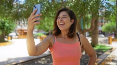 Akıllı telefondan konuşan bir kadın video görüşmesi yapıyor güneşli bir günde şehir parkında duruyor ve yüzünde kocaman bir gülümsemeyle sohbetin tadını çıkarıyor..