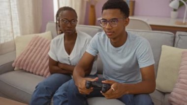 Oğlu evdeki rahat bir oturma odasında kanepede oturup video oyunu oynarken annesi kollarını kavuşturuyor.