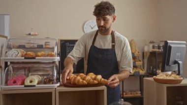 Genç adam pastanede çalışıyor, kruvasanları tezgahın arkasındaki kruvasan tezgahında çeşitli hamur işleri ve fırınlanmış mallarla sıcak bir aydınlatma altında, kapalı bir dükkânda ayarlıyor.