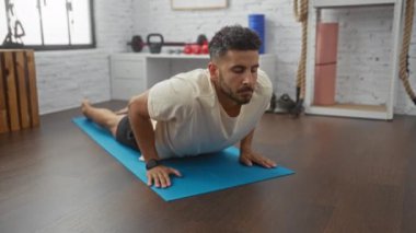 Genç İspanyol bir adam spor merkezinde yoga yapıyor. Geniş, aydınlık bir spor salonunda spor aletleri ve sakin bir atmosferle mavi bir minder üzerinde esneme egzersizi yapıyor..