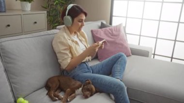 Genç bir kadın rahat bir oturma odasında uyuyan köpeğiyle kanepede otururken telefonuyla oyun oynuyor..
