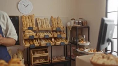 Önlüklü genç adam, arka planda ekmek ve hamur işleriyle dolu raflarla sıcak fırında kahve fincanları tutuyor.