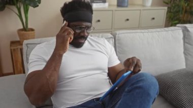 Beyaz tişörtlü Afrikalı Amerikalı adam telefonla konuşuyor ve modern bir oturma odasında tablet kullanıyor..