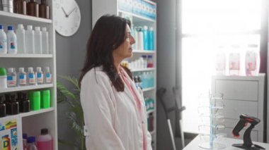 Beyaz önlüklü bir kadın eczanede kolları çapraz duran, şişelerle ve tıbbi ürünlerle dolu raflarla çevrili, profesyonel ve düzenli bir ortam sergileyen