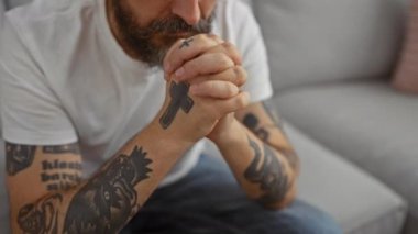 Sakallı ve dövmeli bir adam evinde kanepede otururken dua ediyor.