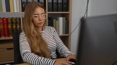 Ofiste çalışan, gözlüklü, uzun sarı saçlı ve çizgili gömlekli bir kadın. Etrafı kitaplık ve belgelerle çevriliyken.