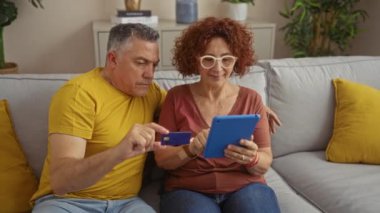 Orta yaşlı bir çift, sıcak bir oturma odasındaki kanepede otururken kredi kartı ve tablet kullanarak evde online alışveriş veya finansal faaliyetler sergiliyor..