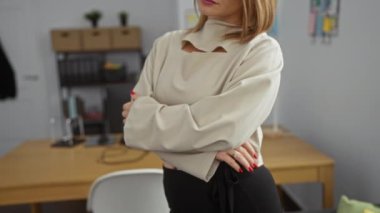 Sarışın bir kadın, arka planda rafları ve masası olan bir ofis ortamında güvenle ayakta duruyor..