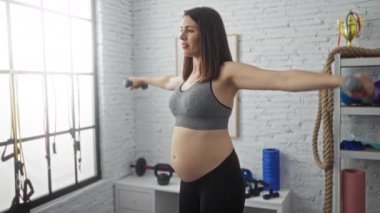 Genç bir İspanyol hamile kadın spor salonunda dambıllarıyla egzersiz yapıyor, spor merkezinde sağlıklı bir hamilelik ve spor merkezine olan bağlılığını gösteriyor..