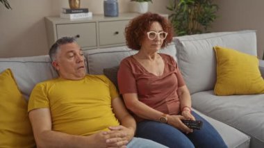 Orta yaşlı bir çift oturma odalarındaki bir kanepede oturup birlikte televizyon seyrediyorlar. Evde sarı yastıklar ve kapalı hava bitkileriyle.