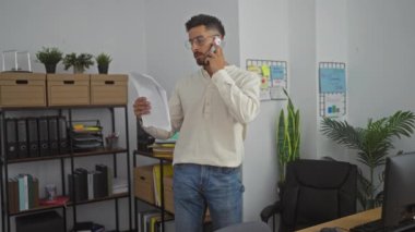 Ofisteki genç İspanyol adam, organize raflar ve duvar çizelgeleri ile çevrili belgeleri tutarken telefonla konuşuyor..