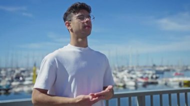 Açık bir günde açık deniz marinasının yanında duran genç İspanyol bir adam ve arkasında mavi gökyüzü olan bir tekne..