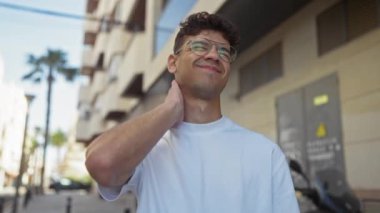 Yakışıklı, İspanyol asıllı, gözlüklü, beyaz gömlekli, gülümseyen ve boynuna dokunan bir adam..