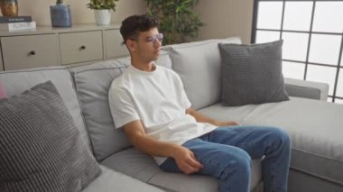 Genç bir adam, modern bir oturma odasında gri bir kanepede oturuyor. Bitkiler ve dekorasyonlarla. Sonra kalkıp odadan çıkıyor ve rahat bir ev ortamı sergiliyor..
