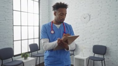 Mavi önlüklü genç bir Afro-Amerikan erkek hemşire bir kliniğin bekleme odasında duruyor, boynunda steteskopla bir not panosuna not yazıyor..