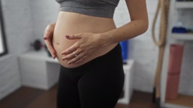Hamile bir kadın spor salonunda karnını tutuyor, spor sutyen ve tayt giyiyor, sağlıklı ve aktif bir annelik hayatı sürüyor..