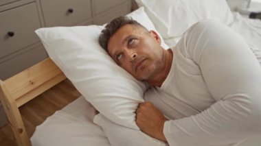 Orta yaşlı bir adam yatak odasında beyaz yastıklarla yatarken yorgun görünüyor ya da uykusuzlukla boğuşuyor.
