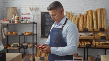 Elinde telefon olan orta yaşlı bir adam fırının içinde duruyor ve arka planda ekmek ve hamur işleri var..