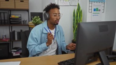 Genç, Afrikalı, Amerikalı, sakallı, kulaklık takan bir adam modern ofis ortamında bir bilgisayarda çalışıyor..