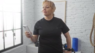 Sarışın, orta yaşlı bir kadın spor salonunda akıllı telefonunu kontrol ediyor. Kapalı bir ortamda çeşitli egzersiz ekipmanlarıyla duruyor..