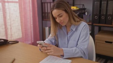 Ofisteki güzel sarışın, beyaz kadın içeride bir masada otururken telefonuna ve evraklarına bakıyor..