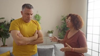 Oturma odasında erkek ve kadın tartışıyor, adam üzgün görünüyor ve kadın tutkulu bir şekilde işaret ediyor, etrafını bitkiler ve ev dekorasyonu sarmış.