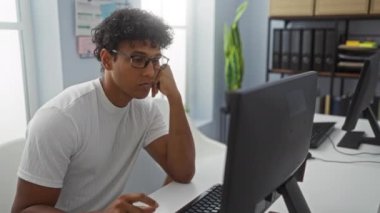 Modern bir ofiste, bilgisayarlarla ve ofis malzemeleriyle çevrili, yorgunluğu ve iş stresini temsil eden, esneyen ve odaklanmış bir ifadeyle çalışan yorgun genç bir adam..