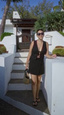 Lanzarote, kanarya adalarında genç bir kadın, siyah elbise ve güneş gözlüğü takıyor, elinde saman bir çanta, beyaz binalar ve yeşilliklerle çevrili, gün ışığında gülümsüyor..