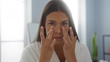 Güzellik salonunda cilt bakım rutini uygulayan bir kadın, beyaz bir bornoz giyiyor, esmer, genç ve çekici, kapalı bir sağlık odasında yüzüne odaklanıyor..