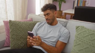 Kanepede oturan İspanyol adam yeşil yastıklı ve pastel desenli rahat bir oturma odasında endişeli bir ifadeyle telefonuna bakıyor.